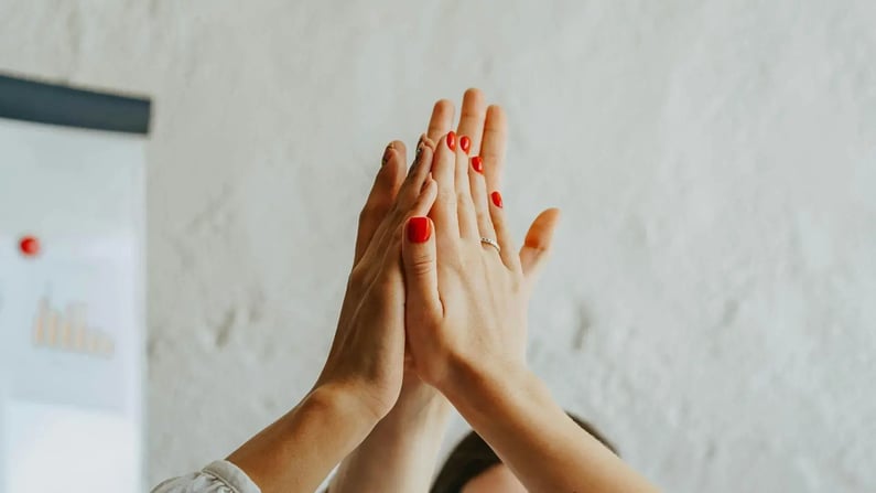 Eine Nahaufnahme zeigt zwei Hände, die in einer High-Five-Geste aufeinandertreffen. Eine Hand hat rote Fingernägel und trägt einen Ring. Die Szene, vor einer weißen Wand, vermittelt Zusammenarbeit und Erfolg.