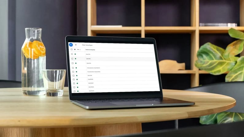 Ein Laptop steht auf einem runden Holztisch in einem Büro. Auf dem Bildschirm ist eine Liste von Google-Gebotsstrategien in deutscher Sprache zu sehen. Links daneben steht eine Wasserkaraffe mit Zitronenscheiben und ein Glas. Im Hintergrund ist ein Regal und eine große Zimmerpflanze zu erkennen