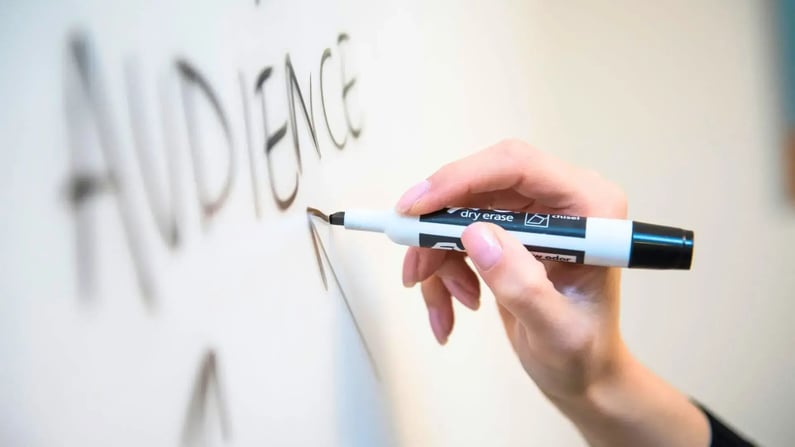 Eine Nahaufnahme zeigt eine Hand mit hellrosa lackierten Fingernägeln, die mit einem schwarzen Whiteboard-Marker das Wort "AUDIENCE" auf eine weiße Tafel schreibt. Der bereits geschriebene Teil des Wortes ist unscharf, während die Hand und der Marker im Fokus liegen.
