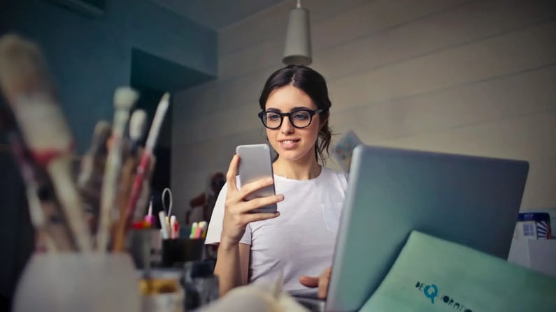 Eine junge, lächelnde Frau mit dunklen Haaren und einer schwarzen Brille sitzt vor einem Laptop an einem Schreibtisch. Sie hält ein Smartphone in der Hand und schaut darauf. Links im Vordergrund sind unscharf Pinsel und Utensilien zu sehen, was auf ein kreatives Umfeld hindeutet.