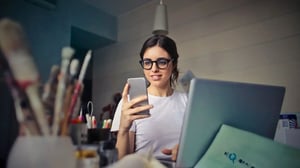 Eine junge, lächelnde Frau mit dunklen Haaren und einer schwarzen Brille sitzt vor einem Laptop an einem Schreibtisch. Sie hält ein Smartphone in der Hand und schaut darauf. Links im Vordergrund sind unscharf Pinsel und Utensilien zu sehen, was auf ein kreatives Umfeld hindeutet.