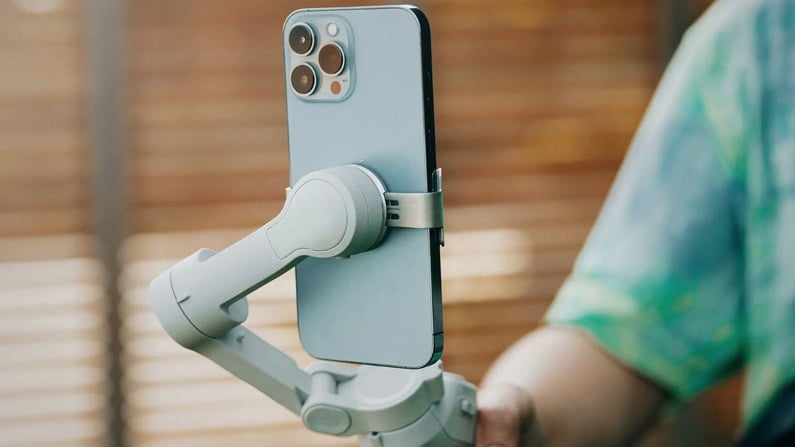 Ein modernes, hellblaues Smartphone mit einem dreifachen Kamerasystem ist in einer weißen Gimbal-Halterung montiert. Das Gerät wird im Freien gehalten, erkennbar an einem unscharfen, braunen Hintergrund (vermutlich Holz) und einem Teil einer grün-türkisen Kleidung rechts.