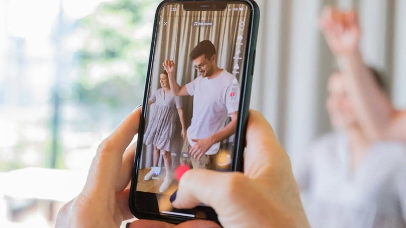 Hände halten ein Smartphone, das eine kurze Video-App zeigt. Auf dem Bildschirm sind eine Frau im Kleid und ein Mann im weißen T-Shirt beim Tanzen zu sehen, wahrscheinlich für TikTok oder Reels. Eine unscharfe Person im Hintergrund hebt ebenfalls die Arme.