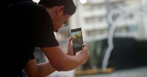 Ein Mann in schwarzem T-Shirt und Brille hält ein Smartphone und blickt auf den Bildschirm. Er fotografiert oder filmt etwas im Freien. Auf dem Display ist eine Person vor einer bunten, gemusterten Wand zu sehen. Der Hintergrund ist unscharf und zeigt eine städtische Umgebung.