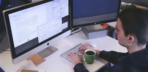 Ein Mann sitzt an einem weißen Schreibtisch und arbeitet an zwei Monitoren. Auf dem linken Bildschirm sind Dokumente und Text zu sehen, der rechte Bildschirm zeigt einen dunklen Hintergrund, möglicherweise ein Spiel oder Code. Er tippt auf einer Tastatur, während eine grüne Tasse Kaffee vor ihm steht.