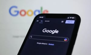 Ein Smartphone im Querformat zeigt die Google-Suchoberfläche im dunklen Modus (Dark Mode). Im Hintergrund ist das klassische, bunte Google-Logo unscharf auf einem größeren Bildschirm zu sehen.